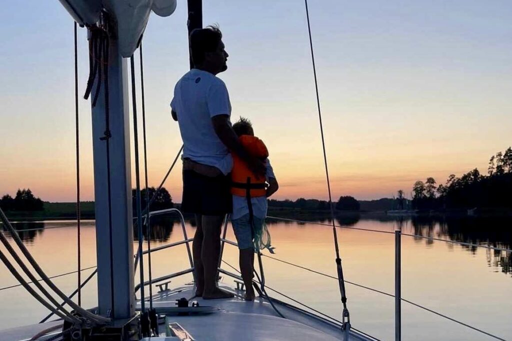 Tata z synem na dziobie jachtu, obserwują zachód słońca w mazurskiej zatoce.