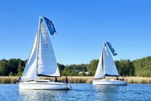 Co to jest żeglarstwo? Mazury atrakcje turystyczne Dwa jachty żaglowe płynące po mazurskim jeziorze w słoneczny dzień, z powiewającymi biało-niebieskimi flagami i zielonym brzegiem w tle.