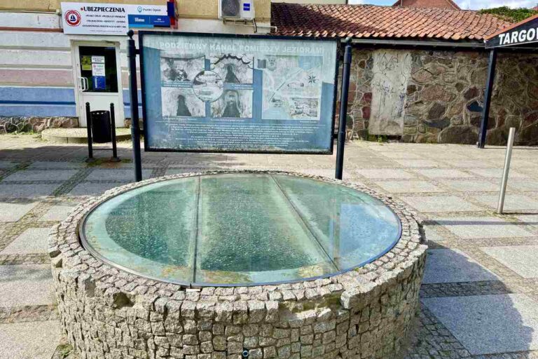 Podziemny kanał między jeziorami w Rynie – szklana tafla i tablica informacyjna.